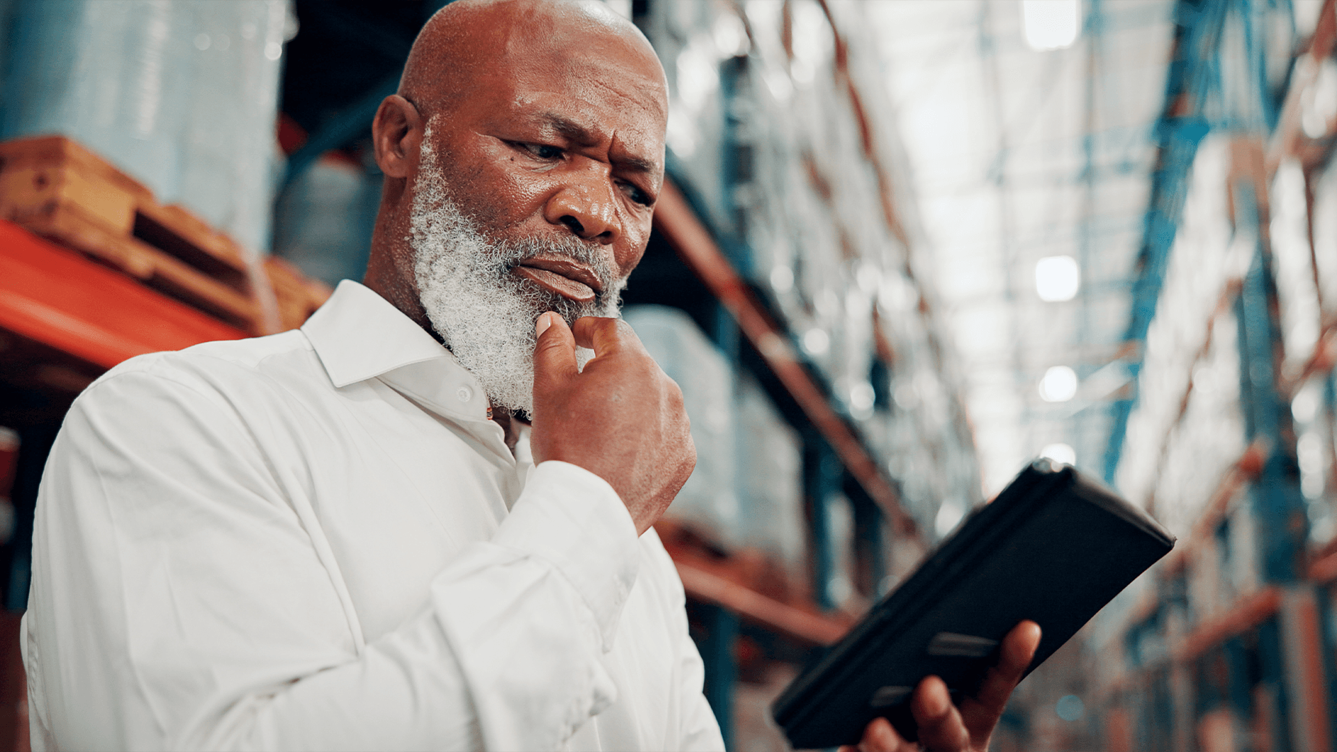 Warehouse manager with a white beard studying a tablet, looking thoughtful, with pallet racking in the background.