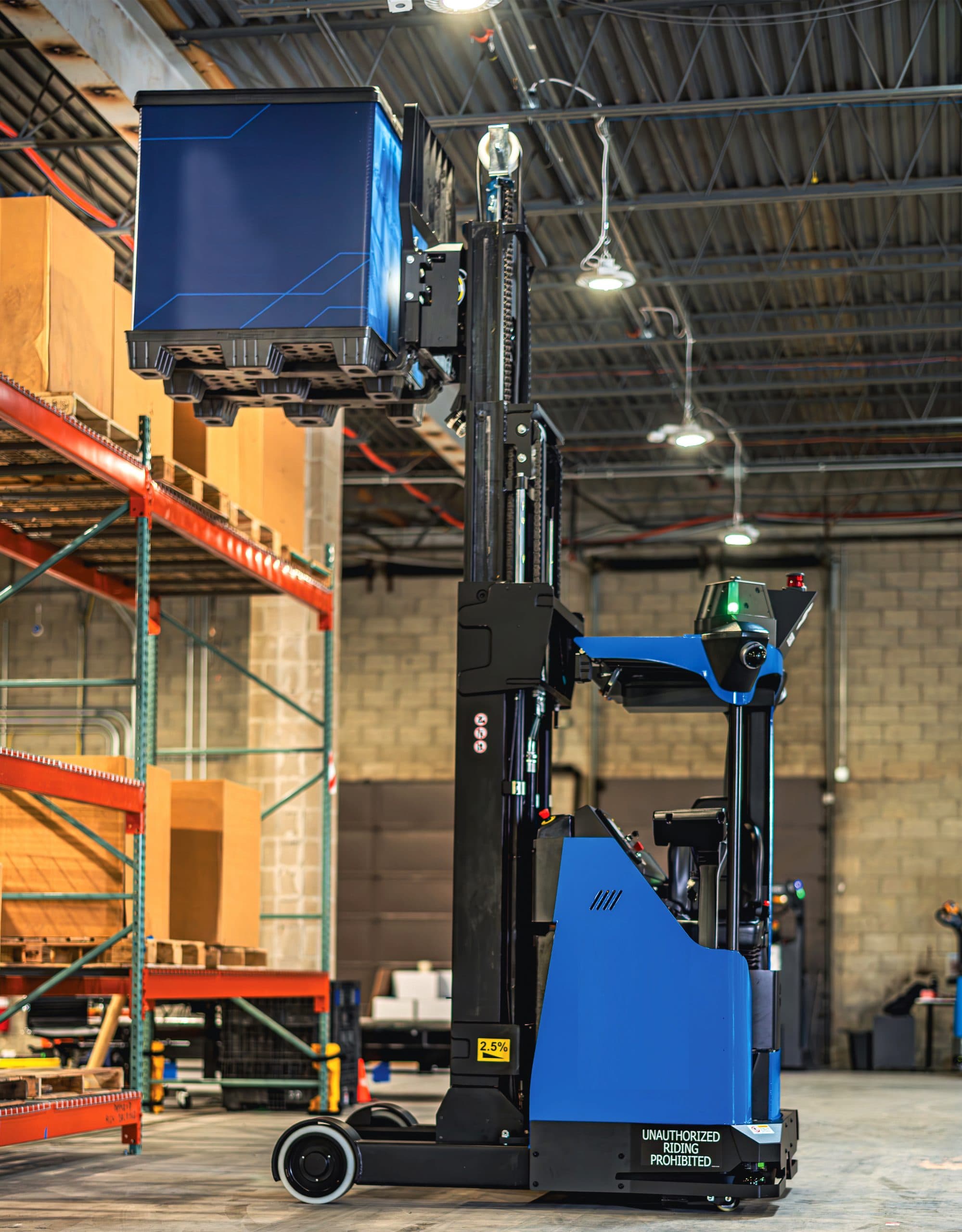 Automated blue and black forklift lifting a large blue container in a warehouse with high ceilings, shelving, and visible safety signage.