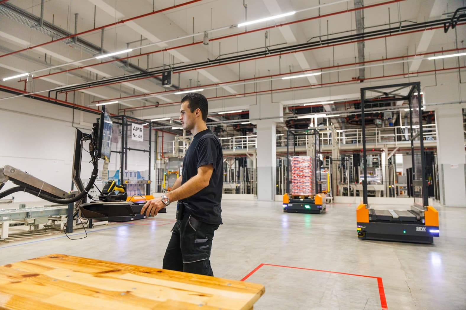 Person operating machinery in a warehouse, holding a control device while monitoring equipment. Automated guided vehicles (AGVs) transport goods in the background. The warehouse features high ceilings with exposed pipes and lighting.
